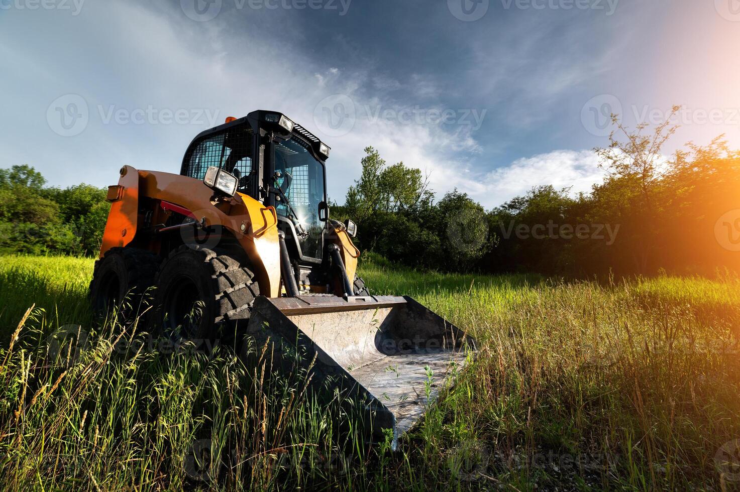small bulldozer stands in green grass against the backdrop of sun rays in a forest belt at sunset photo