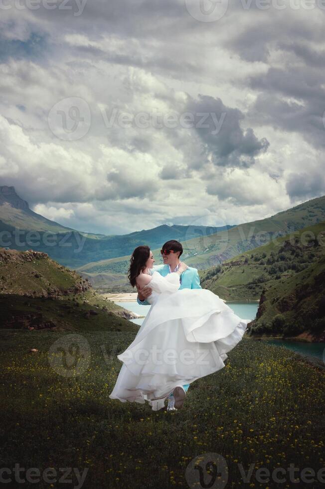 A newlywed couple, a man and a woman, are on a wedding walk in the mountains against the backdrop of rocky mountain peaks and a mountain lake. A man carries his wife in his arms photo