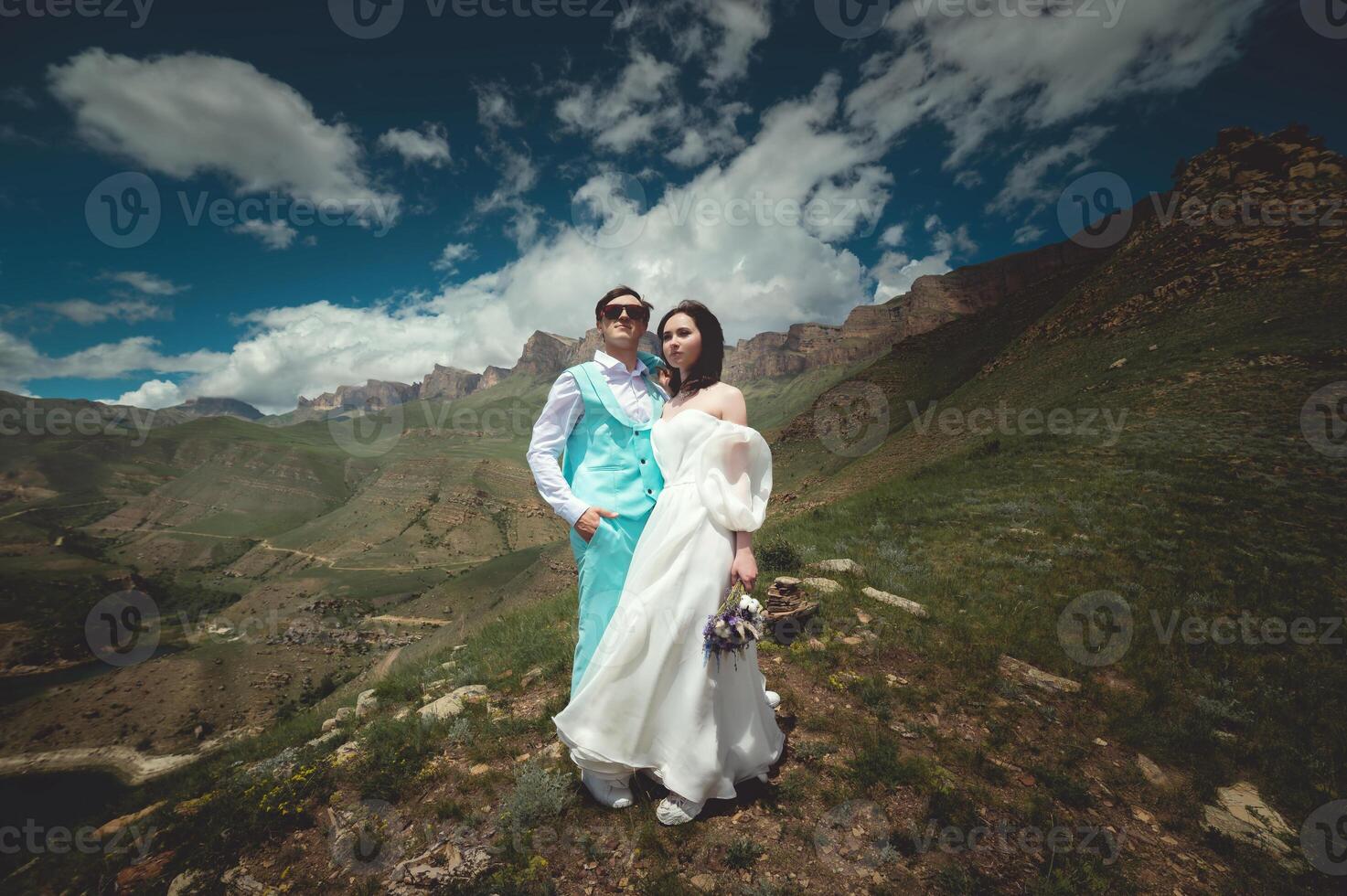 Happy bride and groom hold each other hugging while looking at the sky against the backdrop of hills and mountains. Bride in a fashionable wedding dress with artificial flowers photo