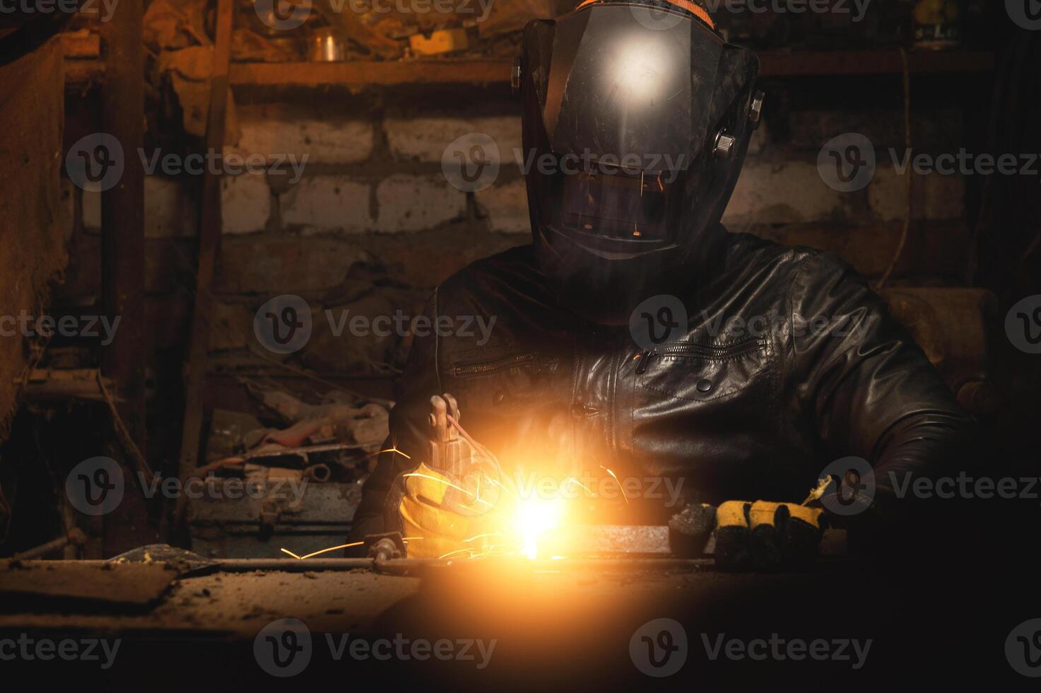 A welder in a protective mask for metal welding and protective gloves in a leather jacket performs welding work in a home garage. Sparks, hot flame from welding. photo