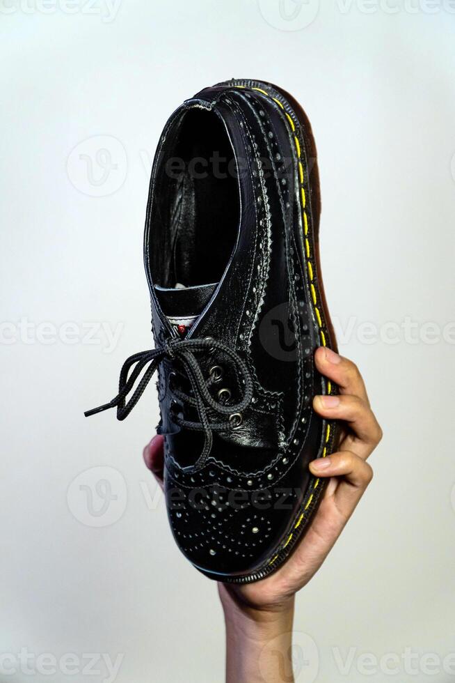 Man's hand holding elegant and shiny full black vintage shoes on a beige background. A jet black brogue wingtip shoe with a rubber outsole is held in a man's hand photo