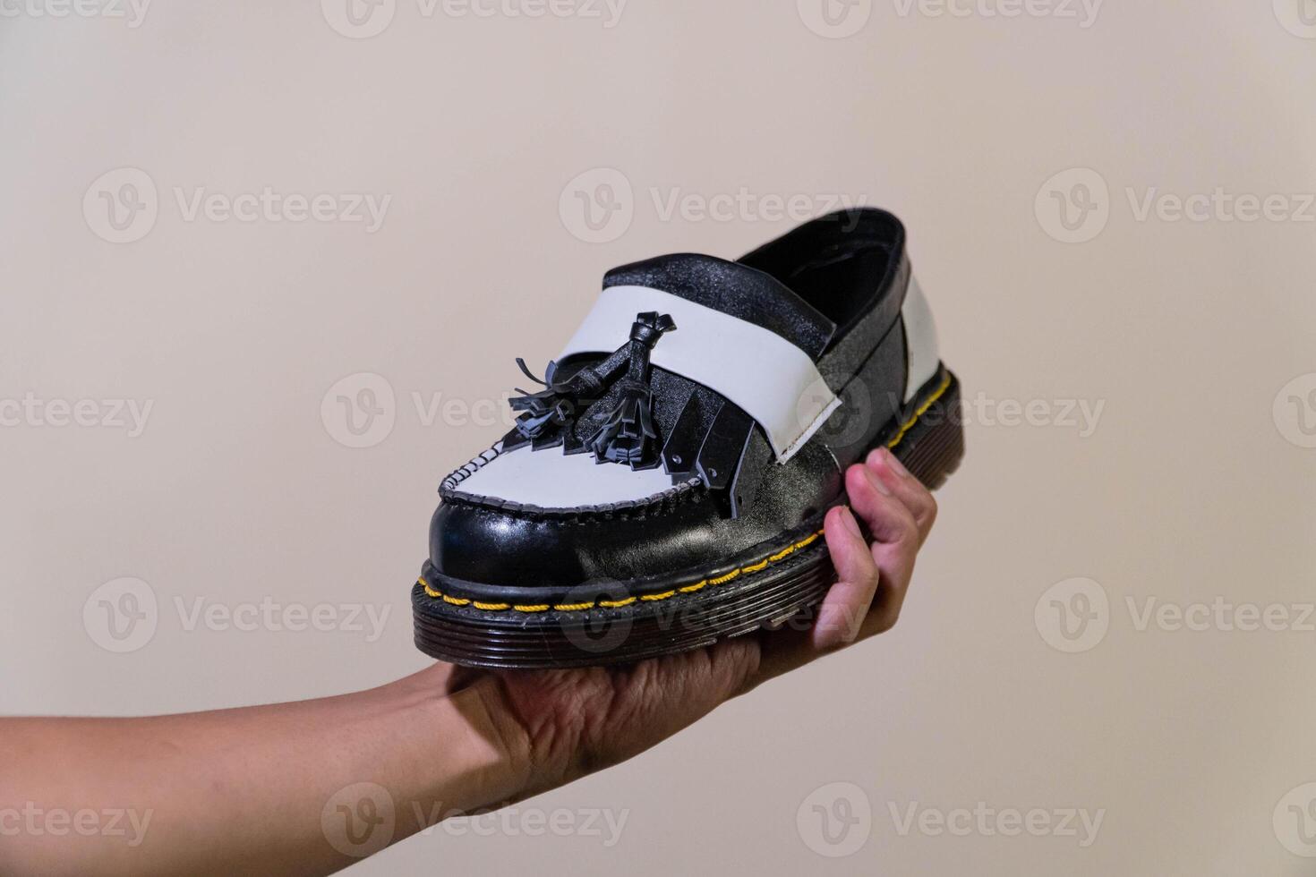 A man's hand holds a black and white tassel shoes with a rubber outsole made from genuine cowhide. Men's hands holding elegant and shiny two tone shoes on a cream background photo