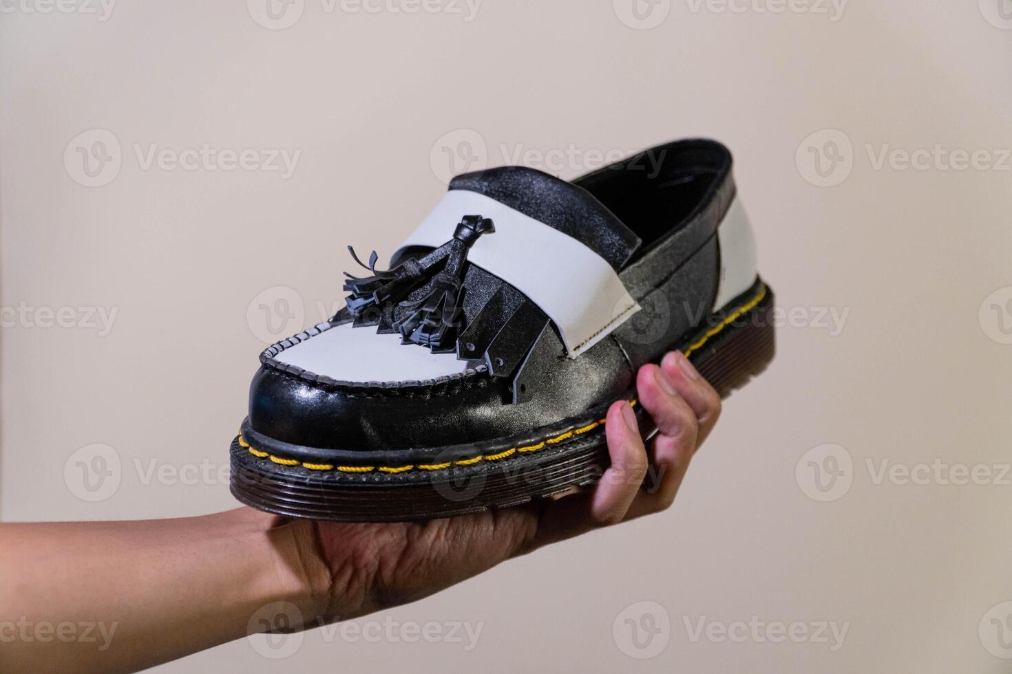 A man's hand holds a black and white tassel shoes with a rubber outsole made from genuine cowhide. Men's hands holding elegant and shiny two tone shoes on a cream background photo