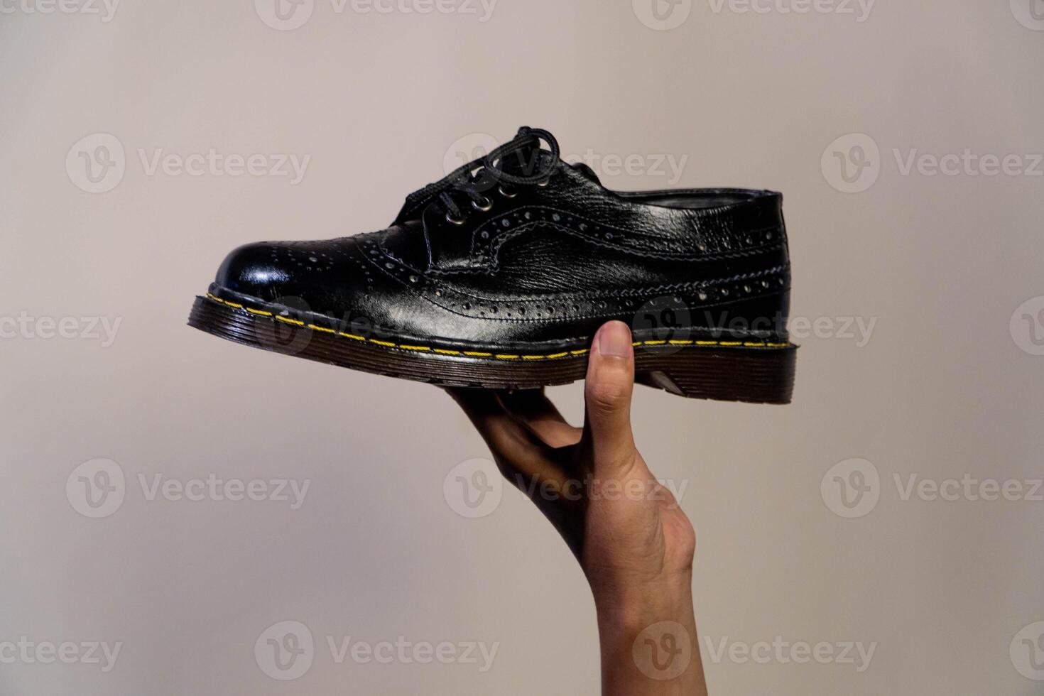 Man's hand holding elegant and shiny full black vintage shoes on a beige background. A jet black brogue wingtip shoe with a rubber outsole is held in a man's hand photo
