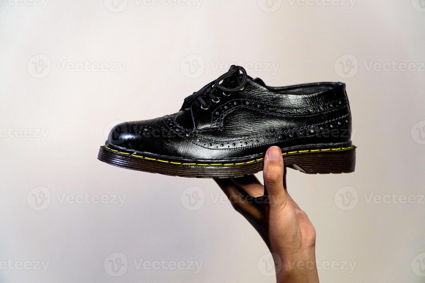 Man's hand holding elegant and shiny full black vintage shoes on a beige background. A jet black brogue wingtip shoe with a rubber outsole is held in a man's hand photo