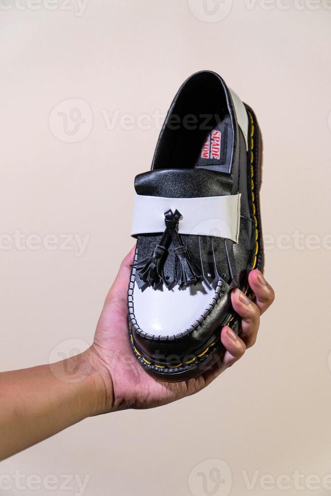 A man's hand holds a black and white tassel shoes with a rubber outsole made from genuine cowhide. Men's hands holding elegant and shiny two tone shoes on a cream background photo
