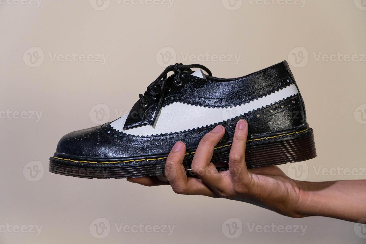 A man's hand holds a black and white brogue wingtip shoe with a rubber outsole made from genuine cowhide. Men's hands holding elegant and shiny two tone shoes on a cream background photo