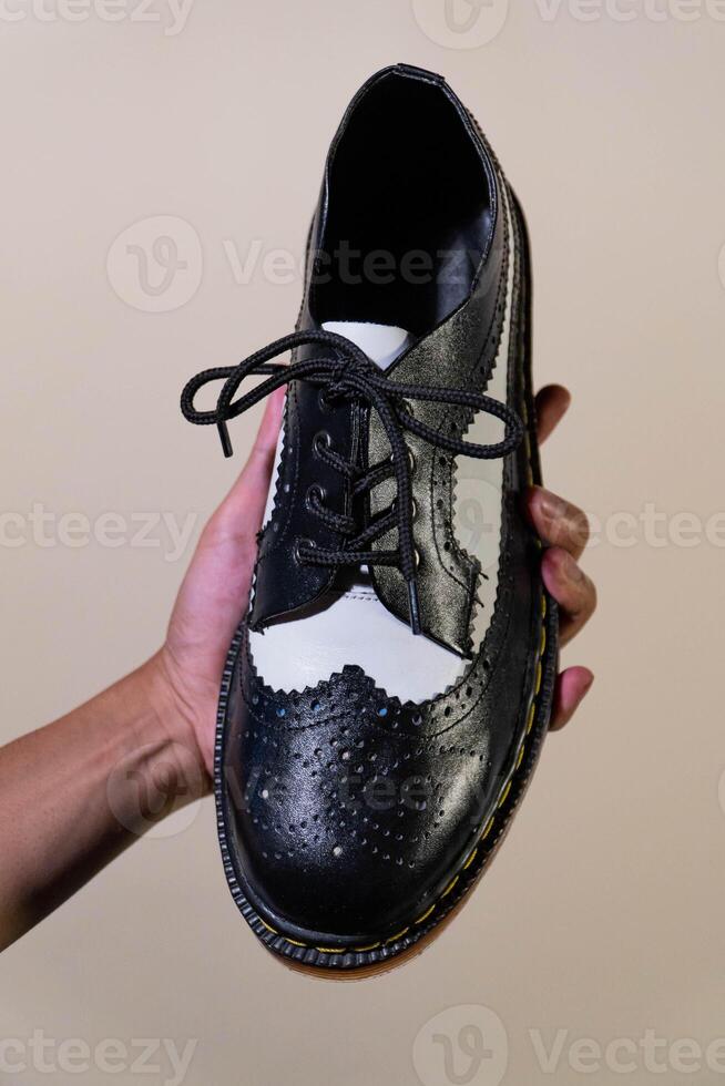A man's hand holds a black and white brogue wingtip shoe with a rubber outsole made from genuine cowhide. Men's hands holding elegant and shiny two tone shoes on a cream background photo