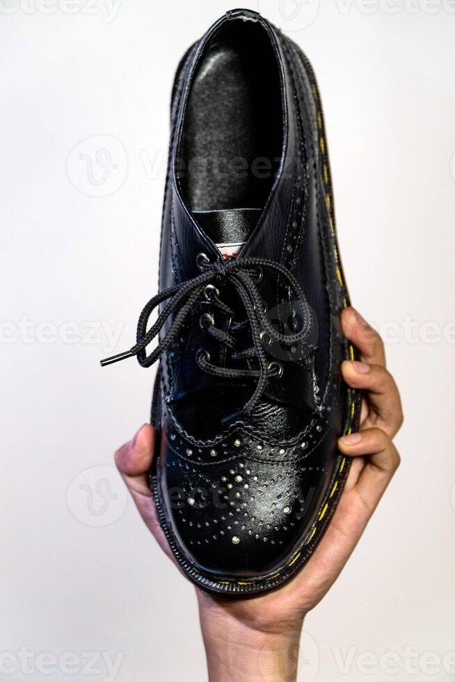 Man's hand holding elegant and shiny full black vintage shoes on a beige background. A jet black brogue wingtip shoe with a rubber outsole is held in a man's hand photo