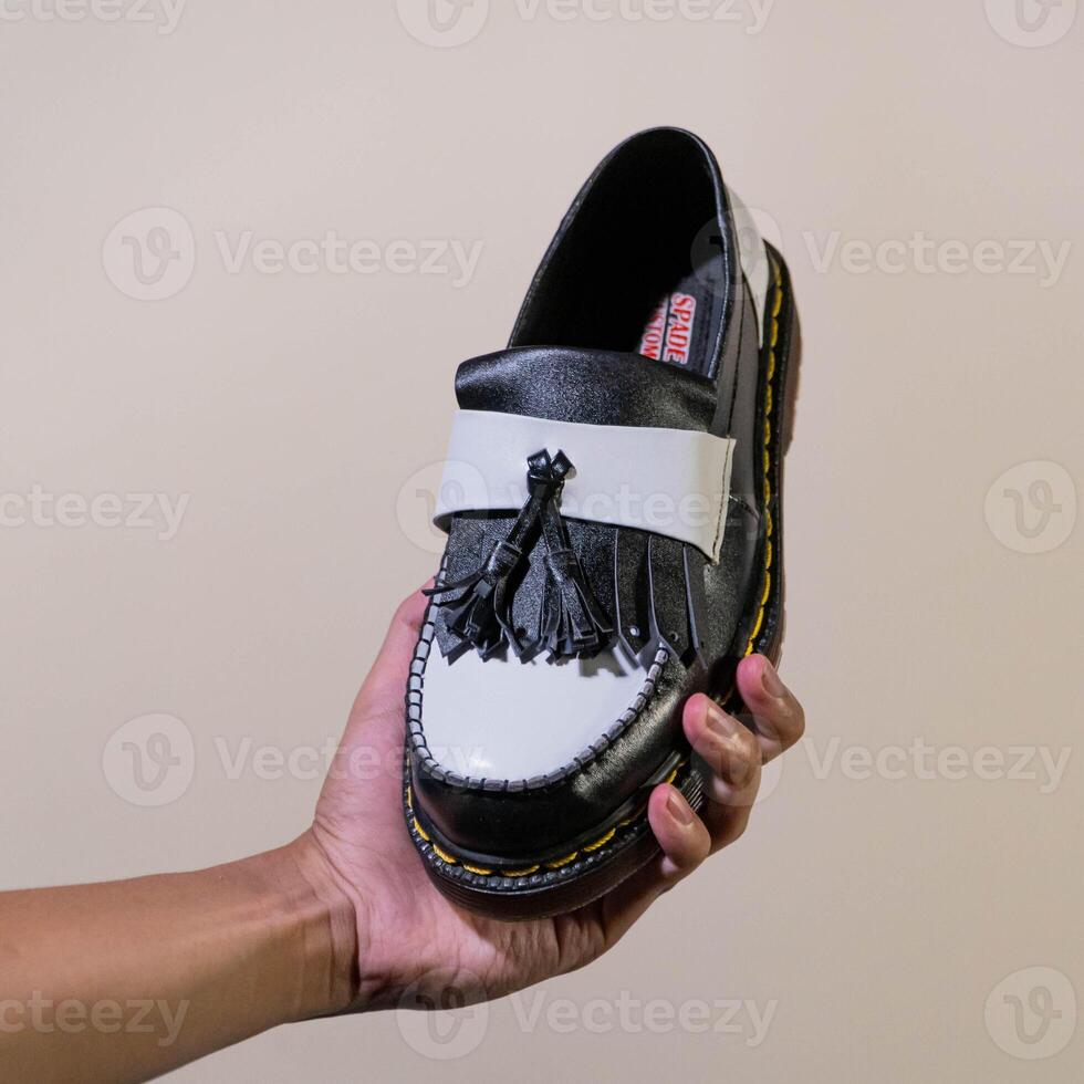 A man's hand holds a black and white tassel shoes with a rubber outsole made from genuine cowhide. Men's hands holding elegant and shiny two tone shoes on a cream background photo