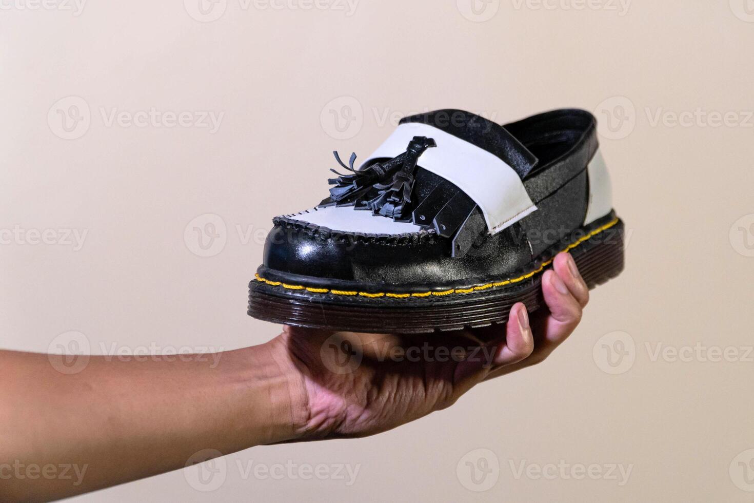 A man's hand holds a black and white tassel shoes with a rubber outsole made from genuine cowhide. Men's hands holding elegant and shiny two tone shoes on a cream background photo