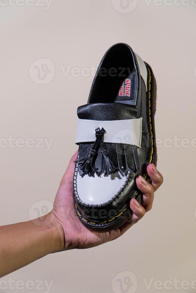 A man's hand holds a black and white tassel shoes with a rubber outsole made from genuine cowhide. Men's hands holding elegant and shiny two tone shoes on a cream background photo