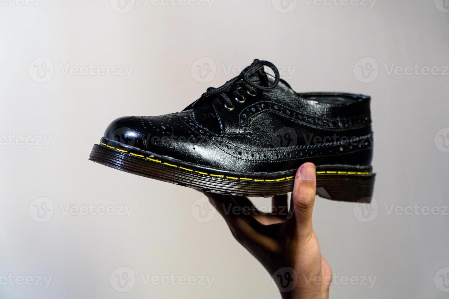 Man's hand holding elegant and shiny full black vintage shoes on a beige background. A jet black brogue wingtip shoe with a rubber outsole is held in a man's hand photo