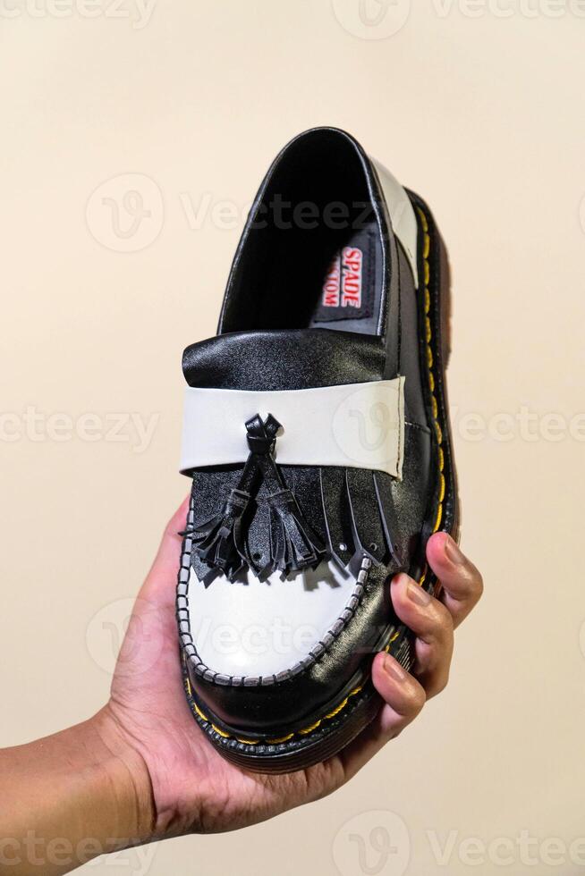 A man's hand holds a black and white tassel shoes with a rubber outsole made from genuine cowhide. Men's hands holding elegant and shiny two tone shoes on a cream background photo