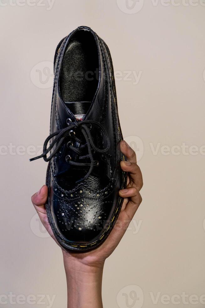 Man's hand holding elegant and shiny full black vintage shoes on a beige background. A jet black brogue wingtip shoe with a rubber outsole is held in a man's hand photo