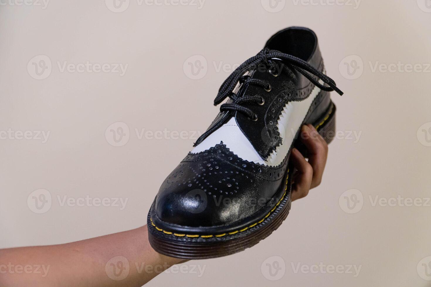 A man's hand holds a black and white brogue wingtip shoe with a rubber outsole made from genuine cowhide. Men's hands holding elegant and shiny two tone shoes on a cream background photo
