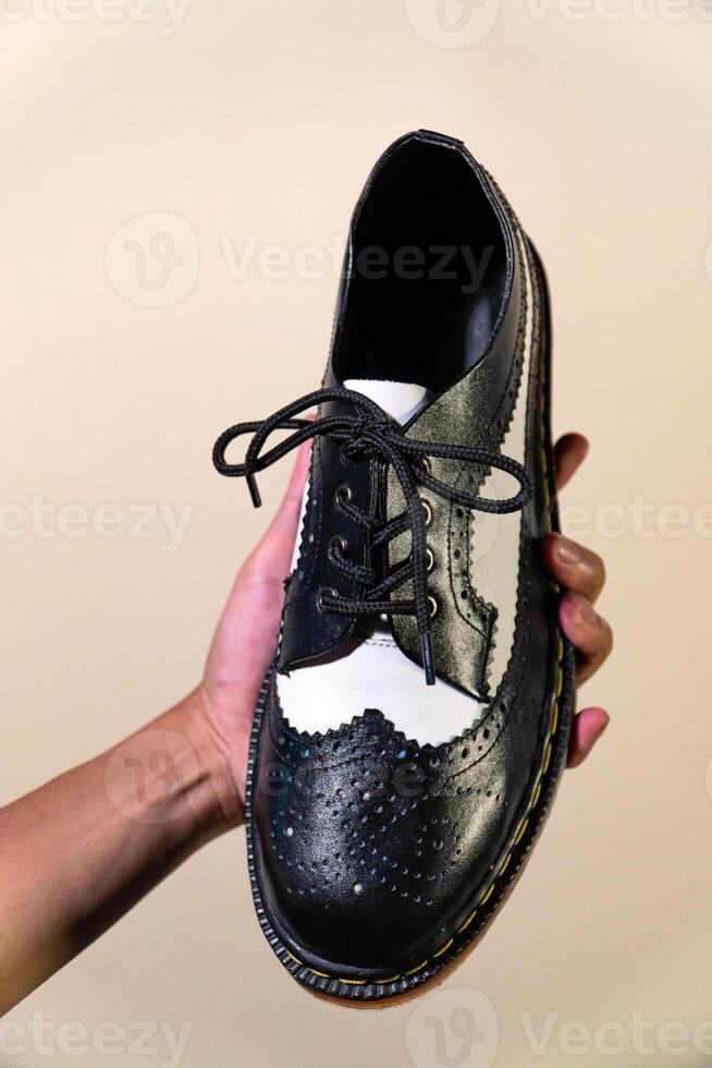 A man's hand holds a black and white brogue wingtip shoe with a rubber outsole made from genuine cowhide. Men's hands holding elegant and shiny two tone shoes on a cream background photo