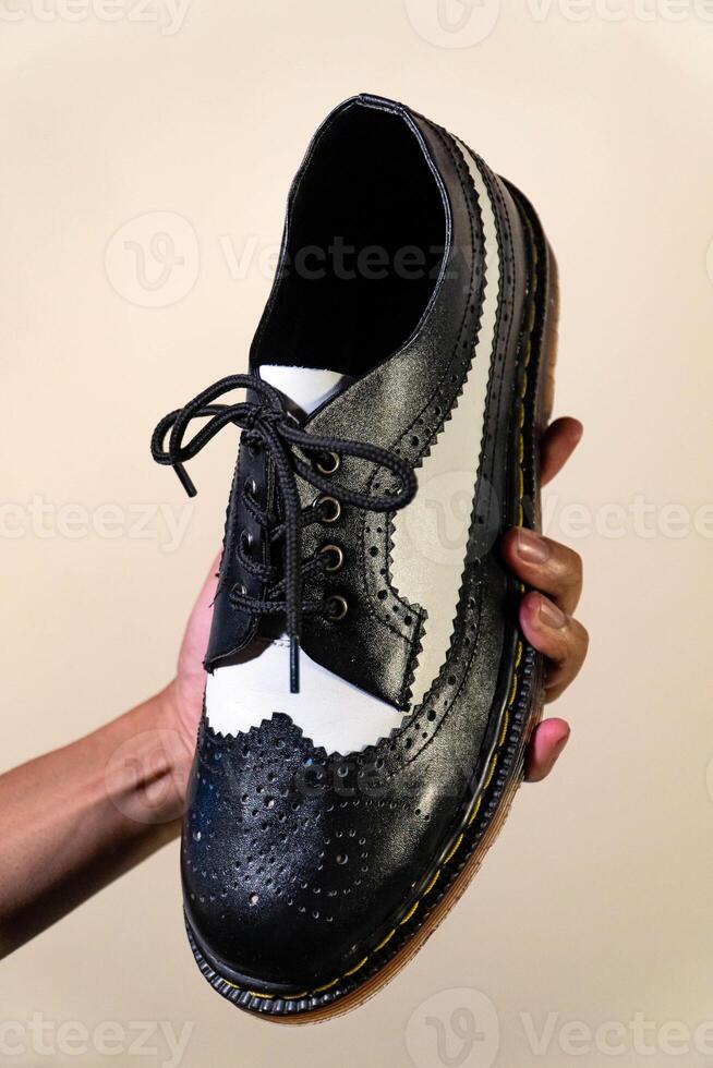 A man's hand holds a black and white brogue wingtip shoe with a rubber outsole made from genuine cowhide. Men's hands holding elegant and shiny two tone shoes on a cream background photo