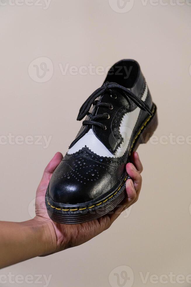 A man's hand holds a black and white brogue wingtip shoe with a rubber outsole made from genuine cowhide. Men's hands holding elegant and shiny two tone shoes on a cream background photo