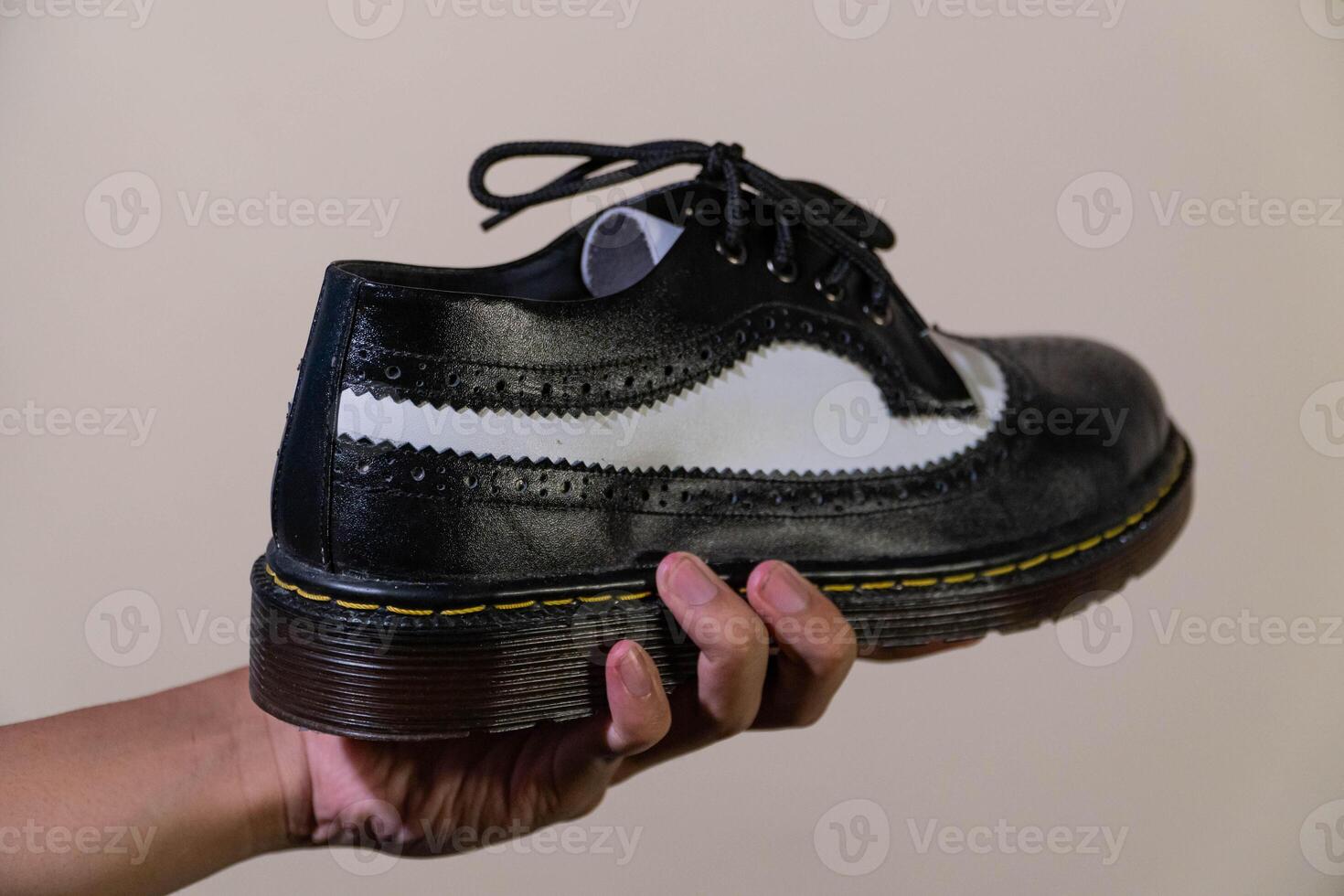 A man's hand holds a black and white brogue wingtip shoe with a rubber outsole made from genuine cowhide. Men's hands holding elegant and shiny two tone shoes on a cream background photo
