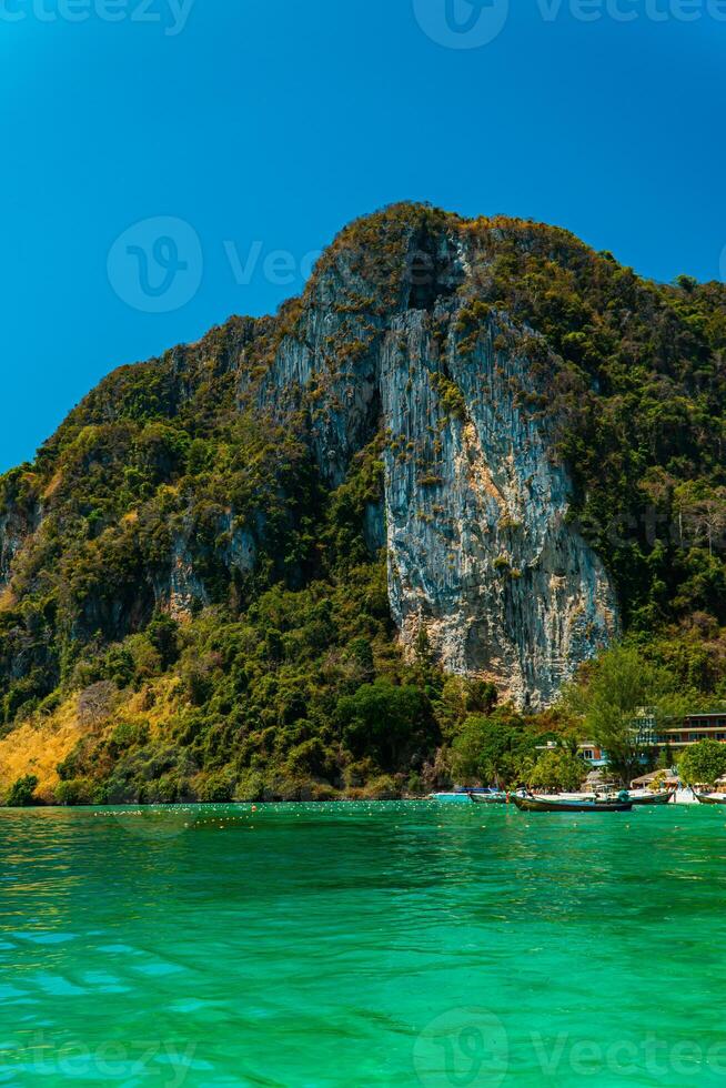 Large rock cliff covered in green trees with the sea in front of it on a sunny day. The view of large rock cliffs and vast ocean is a tourist icon for tropical islands in Krabi, Thailand photo