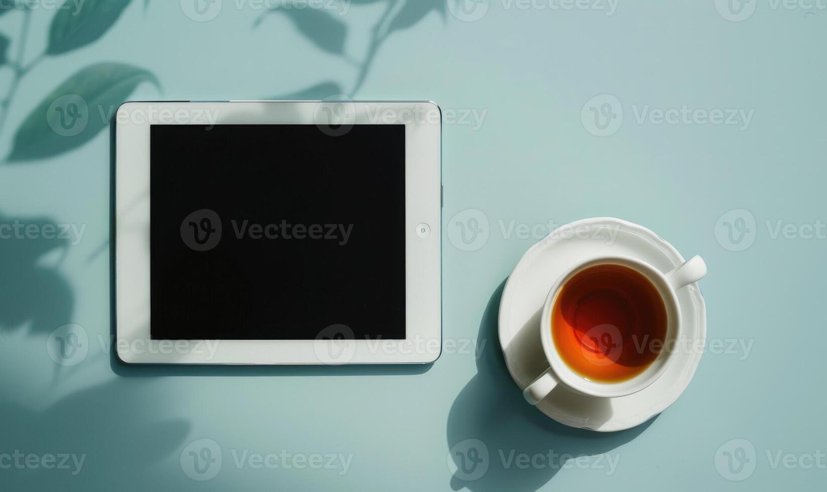 Tablet on a light blue table with a cup of tea on the right, shadow from window, top view photo