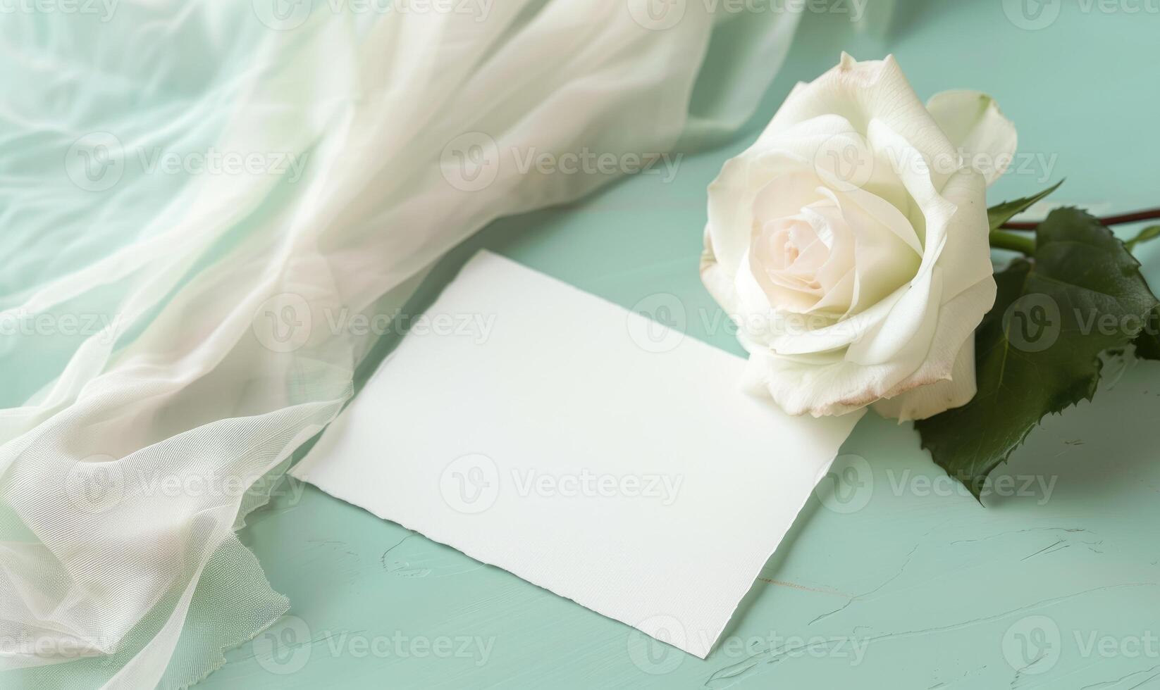 White blank card on a pastel mint green table, with a piece of chiffon fabric and a single white rose photo