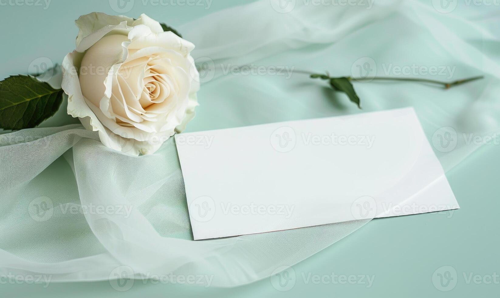 White blank card on a pastel mint green table, with a piece of chiffon fabric and a single white rose photo