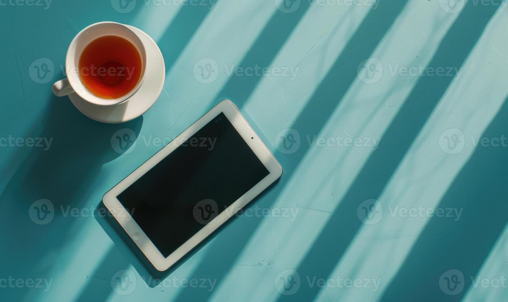 Tablet on a light blue table with a cup of tea on the right, shadow from window, top view photo