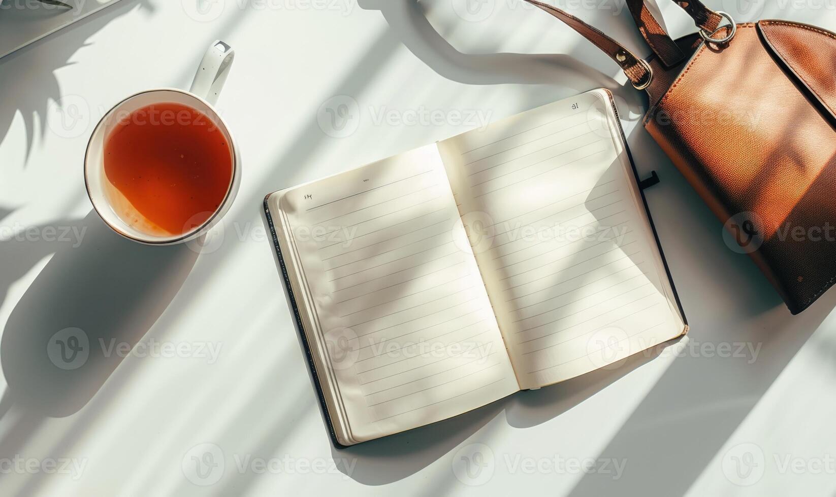 Closed notebook on a light gray table with a cup of tea on the left and a handbag on the right photo