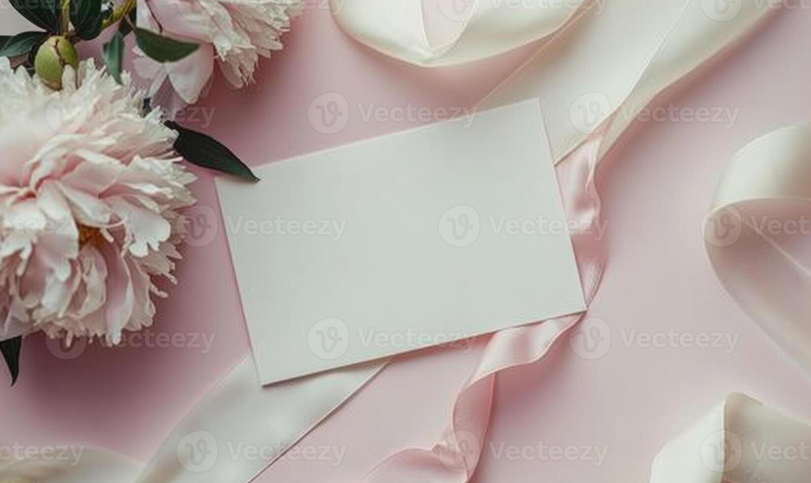 White blank card on a pastel rose pink table, with a piece of satin ribbon and a single peony flower photo