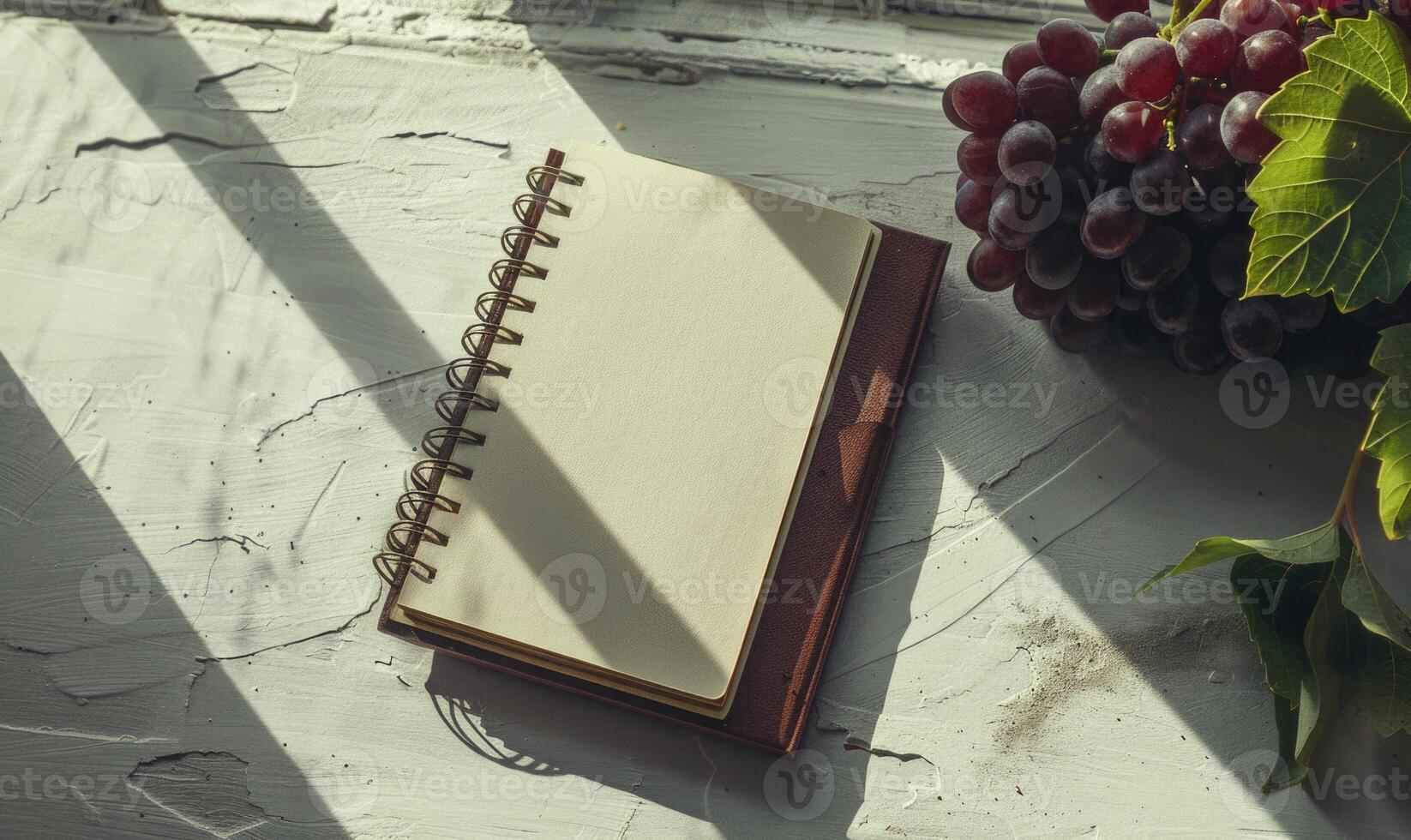 Closed notebook on a light gray table with a bunch of grapes on the right photo