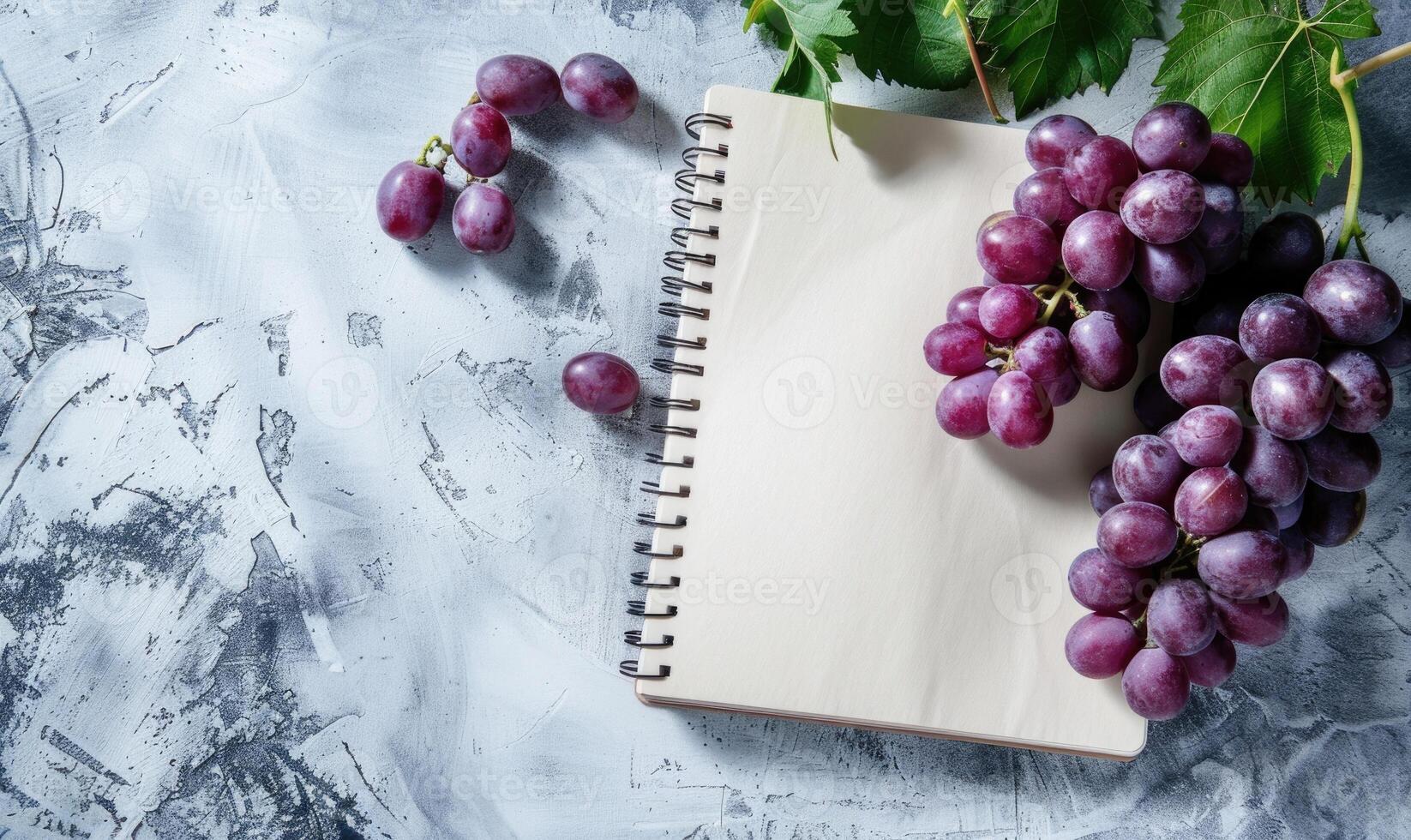 Closed notebook on a light gray table with a bunch of grapes on the right photo
