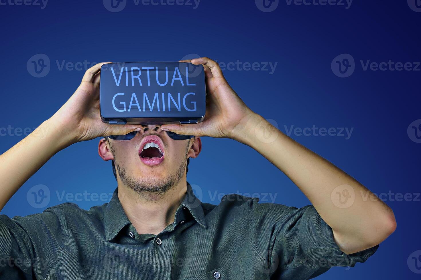 Young man in shirt enjoys and amazed by virtual reality glasses with virtual gaming inscription on them in front of blue background photo
