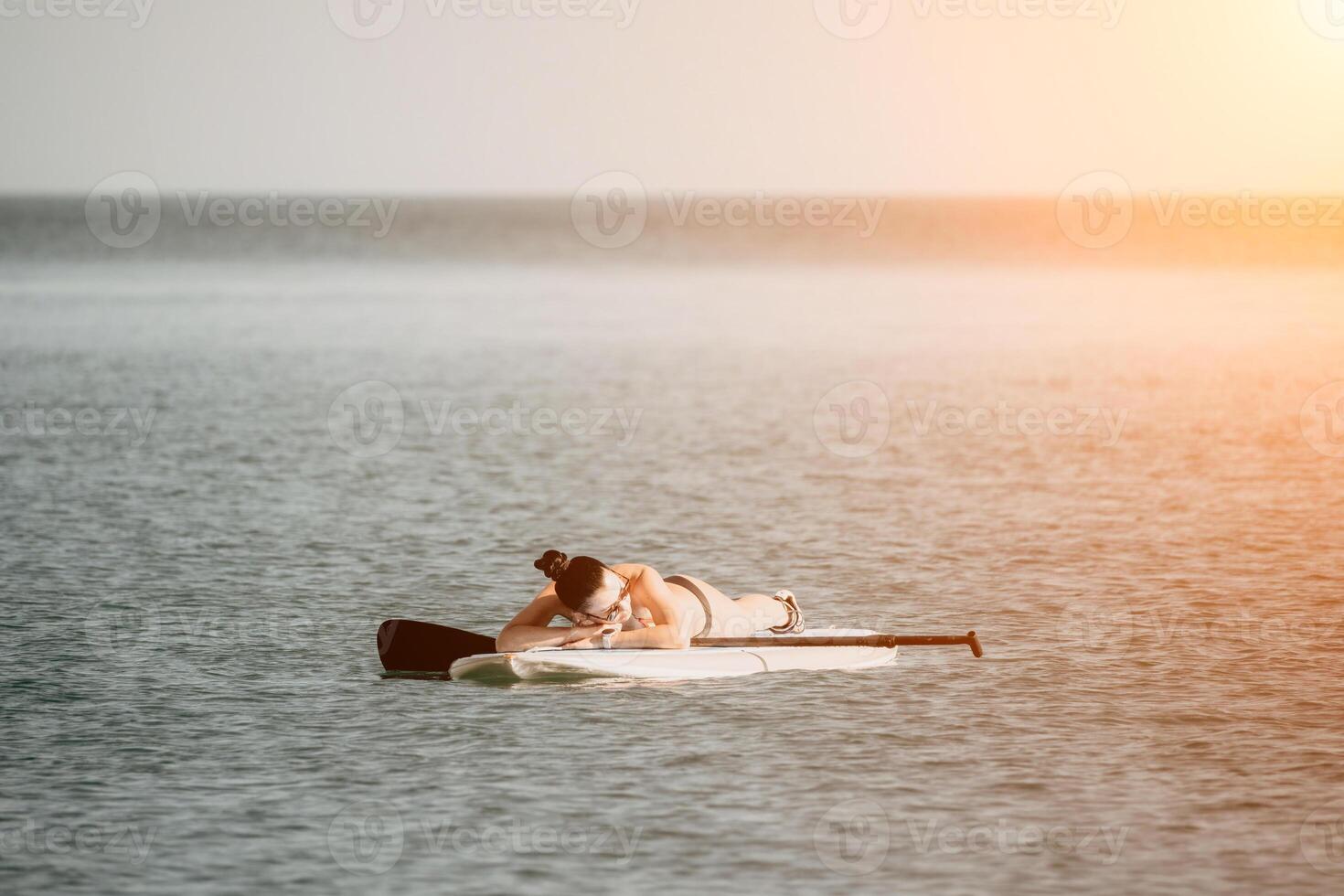 Sea woman sup. Silhouette of happy middle aged tanned woman in rainbow bikini, surfing on SUP board, confident paddling through water surface. Idyllic sunset. Active lifestyle at sea or river. photo