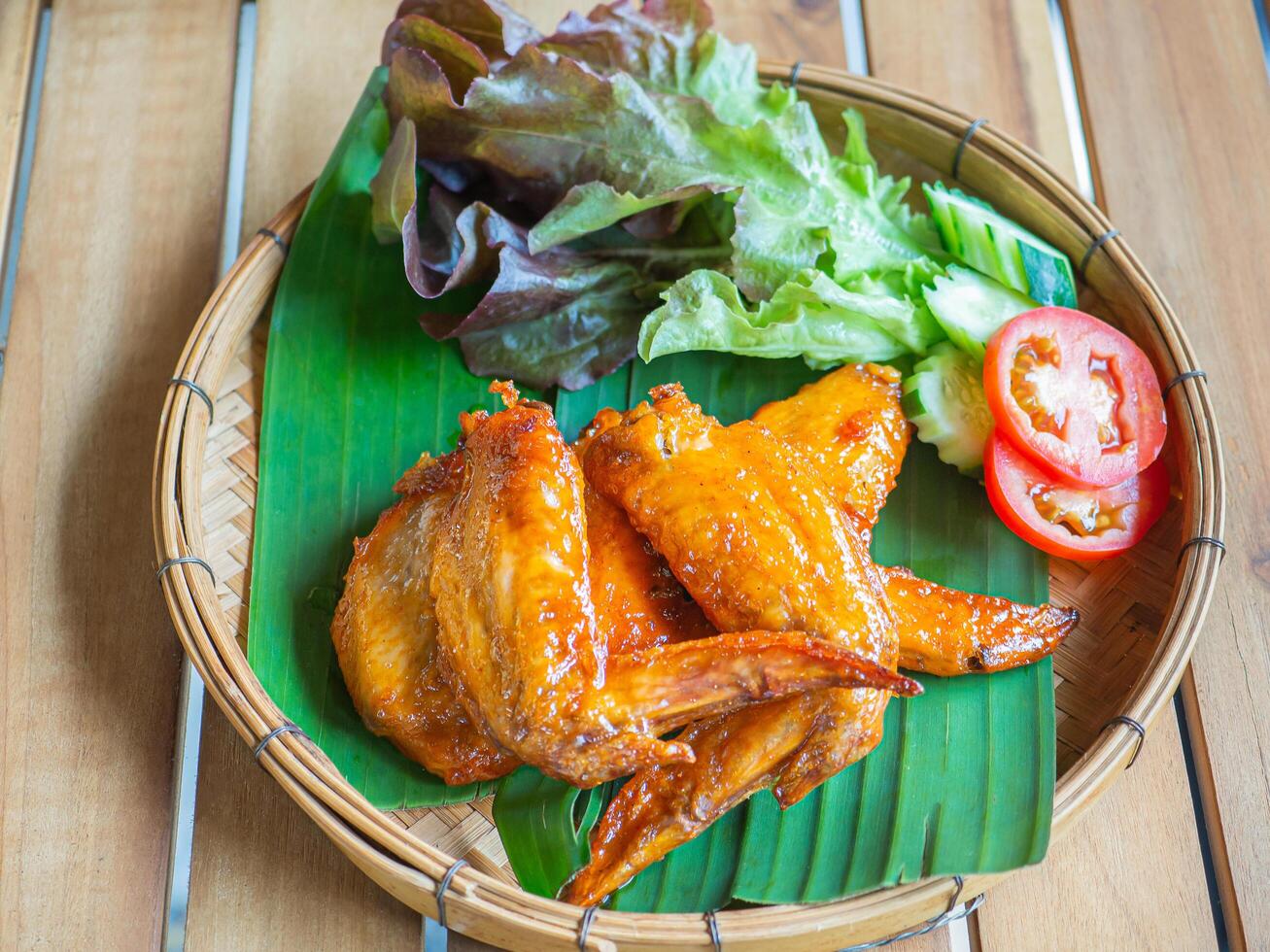 Chicken wing spicy, tomatoes, and lettuce on a banana leaf placed in a bamboo tray. Close-up photo