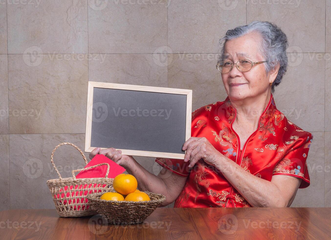 Senior woman wearing a traditional cheongsam qipao dress holding a blackboard and looking at the camera while sitting in the studio with four tangerines and an Ang Pao in a basket. Chinese New Year photo