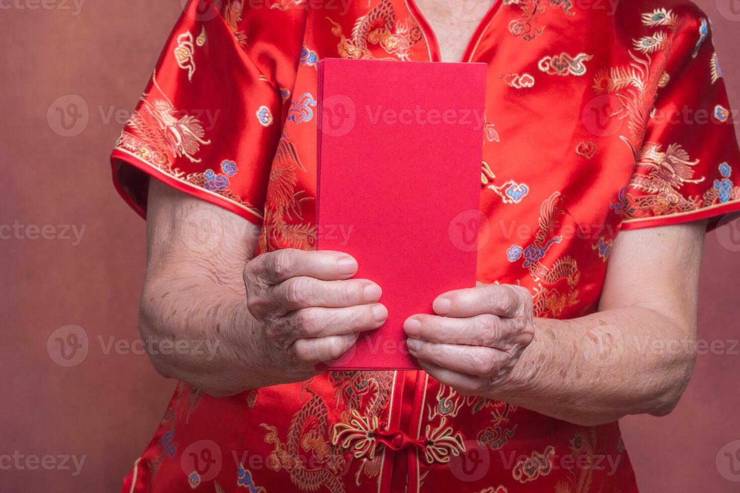Hands of a senior woman holding red envelopes or Angpao and wearing a traditional cheongsam qipao dress while standing in the studio with a vintage background. Close-up photo. Chinese New Year photo