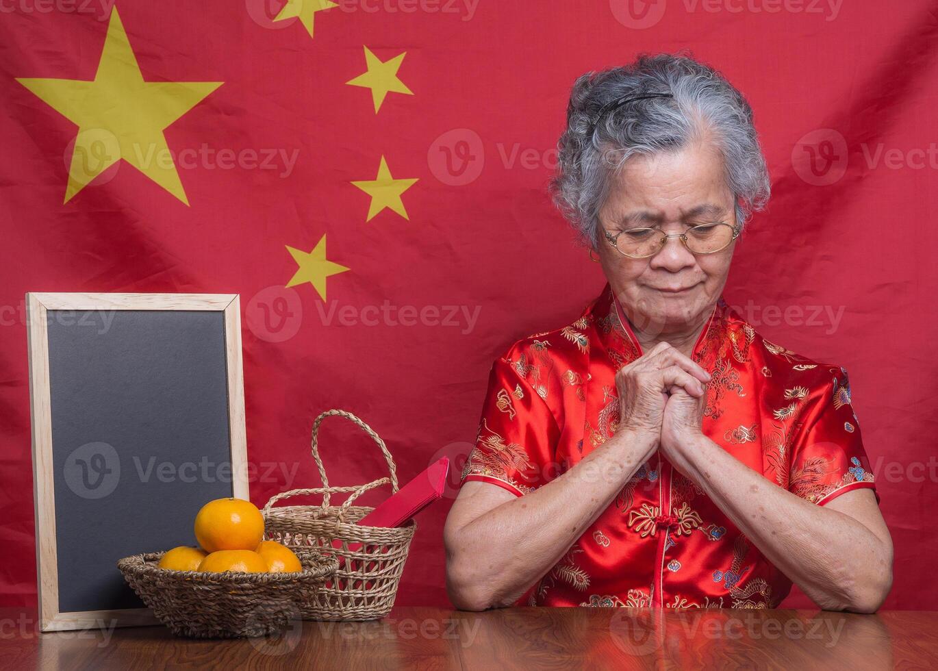 Senior woman wearing a traditional cheongsam qipao dress praying while sitting in the studio with four tangerines in a basket, Aug pow, and a blackboard with a China flag background photo