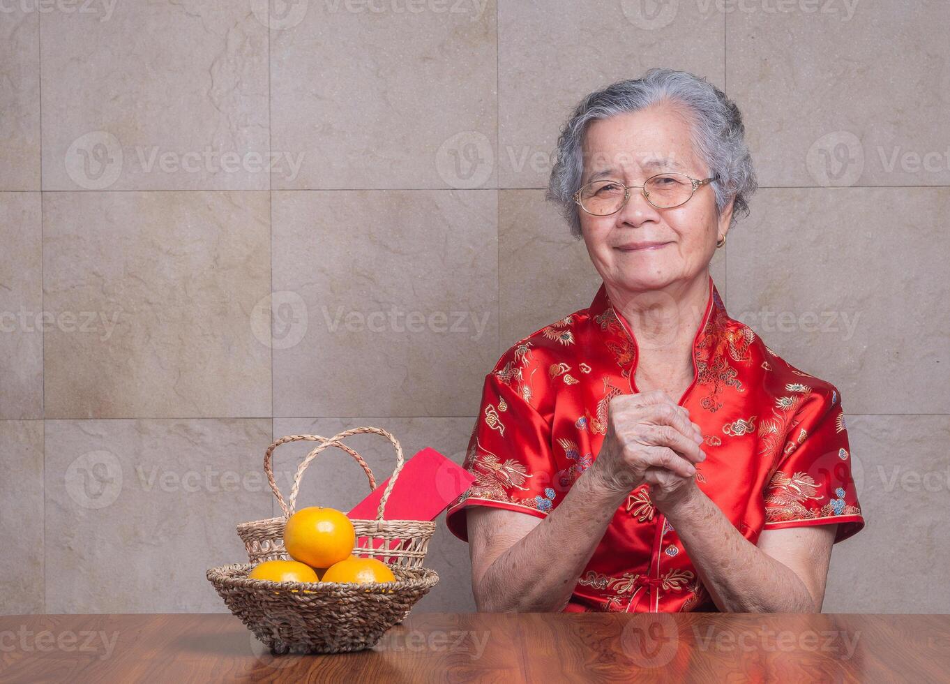 Elderly Asian woman wearing a traditional cheongsam qipao dress looking at the camera while sitting in the studio with four tangerines and an Ang Pao in a basket. Chinese New Year photo