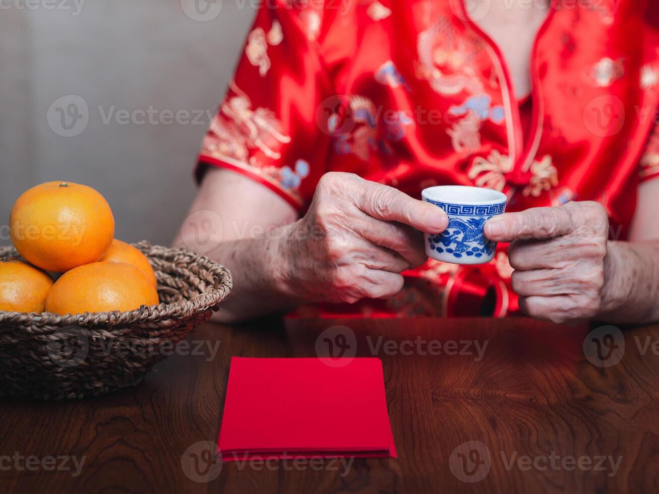 Hands of a senior woman wearing a traditional cheongsam qipao dress holding a teacup with red envelopes or Ang Pao and four tangerines in a basket placed on a wooden table. photo
