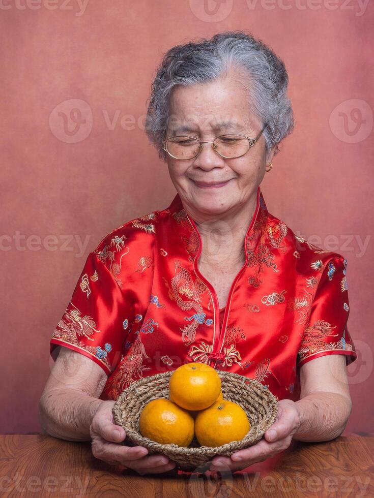 Elderly Asian woman wearing a traditional cheongsam qipao dress holding and looking at a basket with four tangerines while sitting in the studio. Concept of aged people and Chinese New Year photo