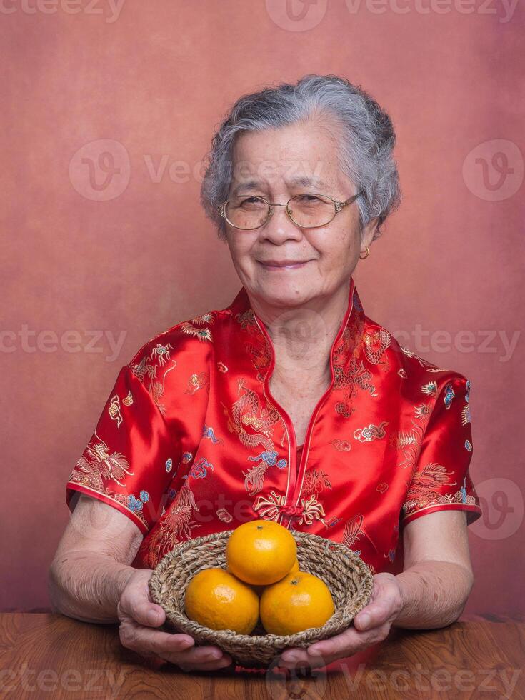 Elderly Asian woman wearing a traditional cheongsam qipao dress holding a basket with four tangerines and looking at the camera while sitting in the studio. Concept of Chinese New Year photo