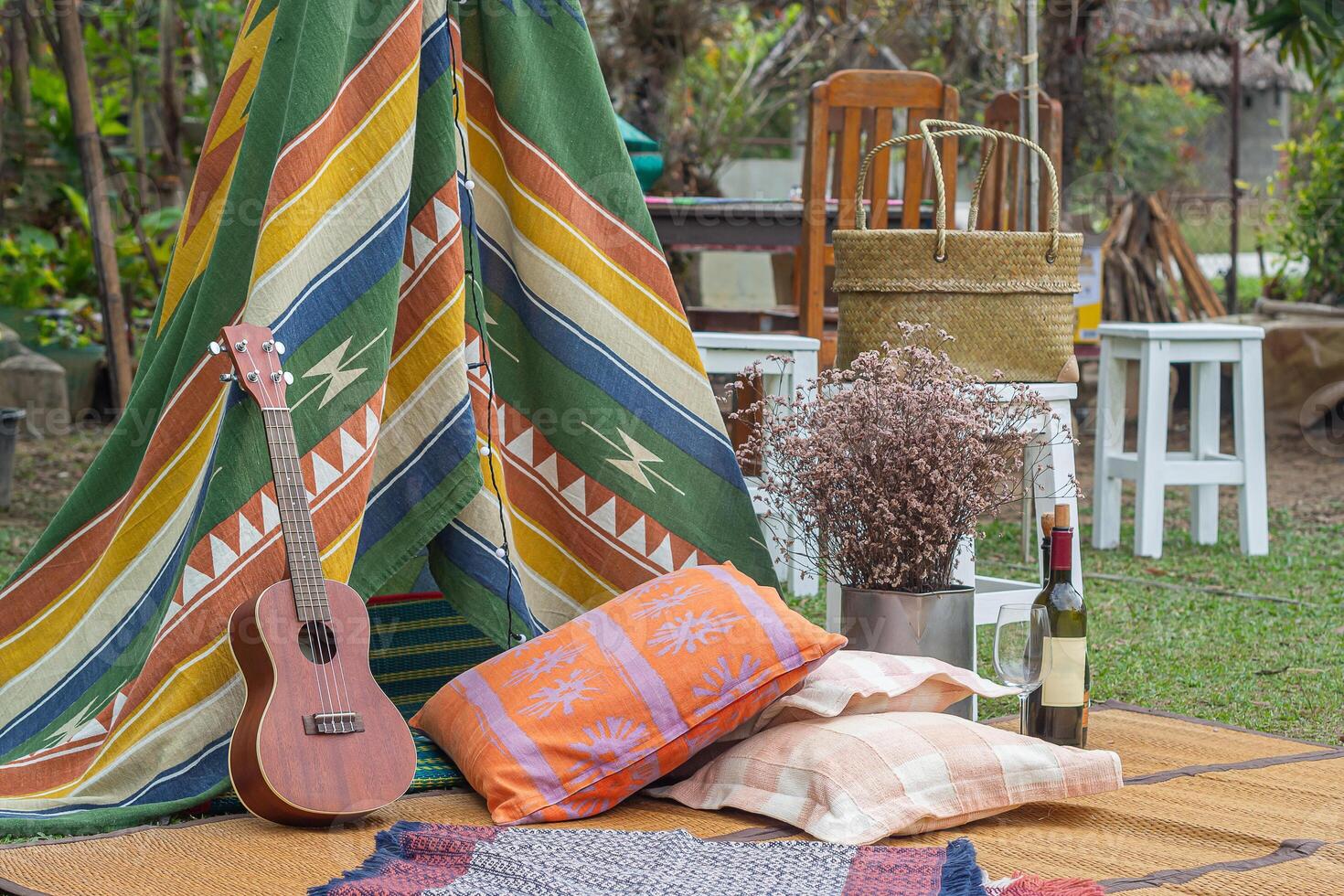 Outdoors area decorated in a Bohemian style with a tent from colorful fabrics, and decorated with the ukulele, pillow, cloth, flower dry, vase, mat, glass, and wine bottle. Concept of relaxation photo