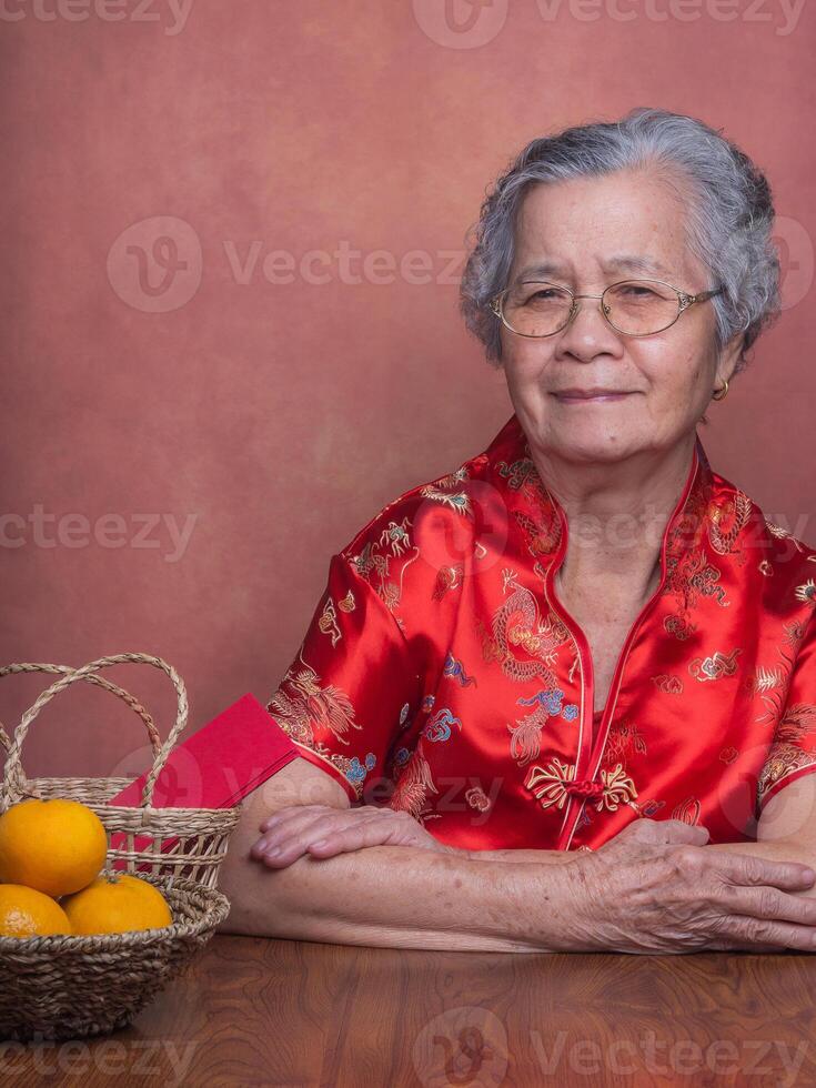 Elderly Asian woman wearing a traditional cheongsam qipao dress looking at the camera while sitting in the studio with four tangerines and an Ang Pao in a basket. Chinese New Year photo