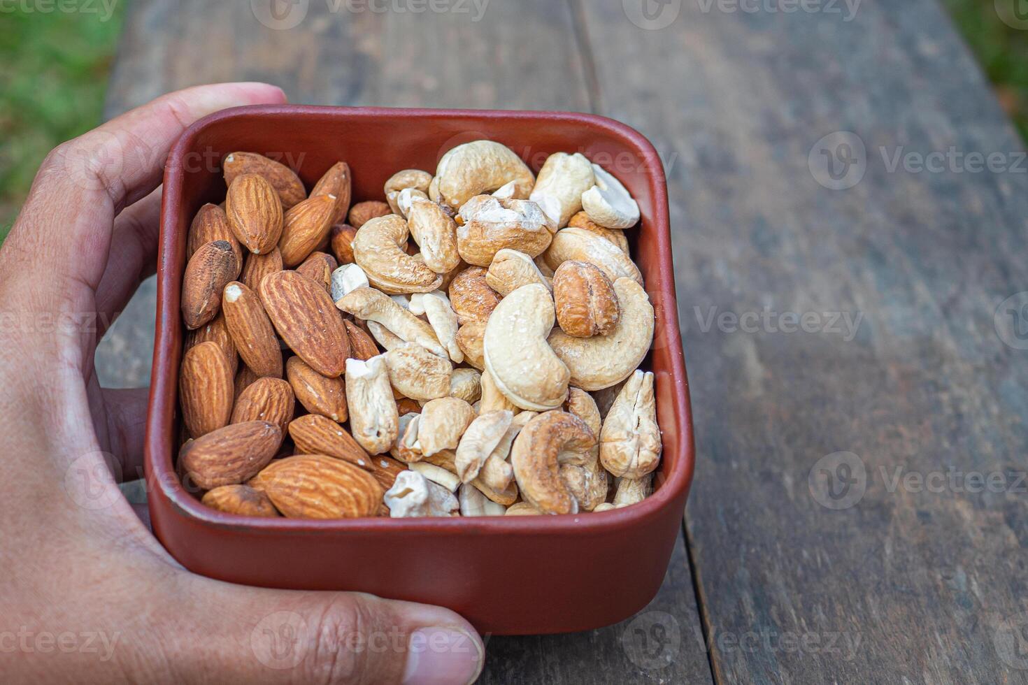 Hand holding of a brown bowl with almonds and cashew nuts on a wooden table. Side view. Space for text. Concept of healthy fruit and snack photo