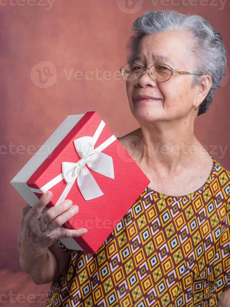 Portrait of an elderly Asian woman holding a red gift box and looking at the camera while standing in the studio. Concept of the festival, anniversary and aged people photo