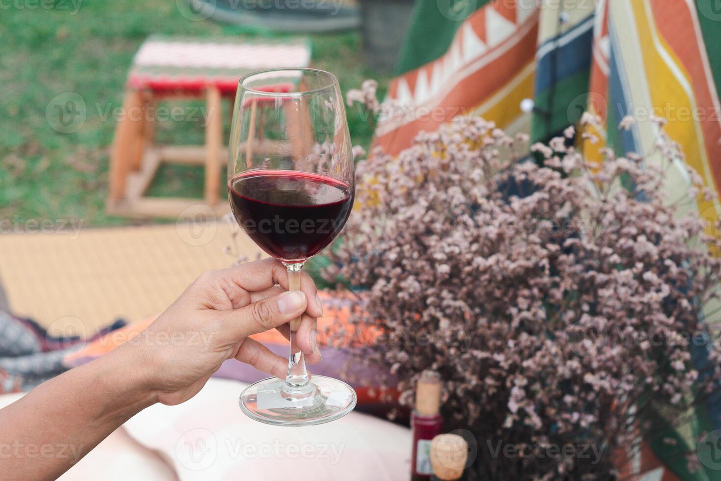 Hand holding of a glass of red wine with a bohemian tent from the colorful fabric and flower background. Concept of relaxation photo