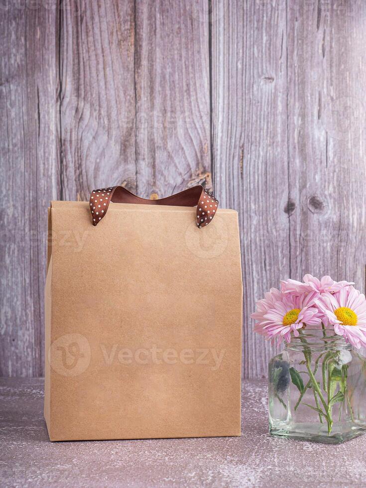 Brown paper bag packaging with ribbon handles placed on a gray stone background with a wooden wall. Side view with copy space for text photo