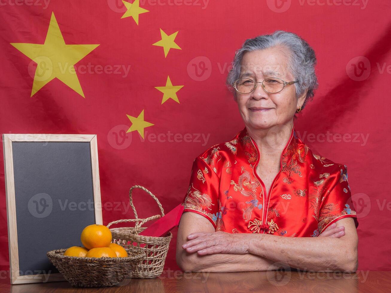 Senior woman wearing a traditional cheongsam qipao dress looking at the camera while sitting in the studio with four tangerines, an Ang Pao in a basket, and a blackboard with a China flag background photo